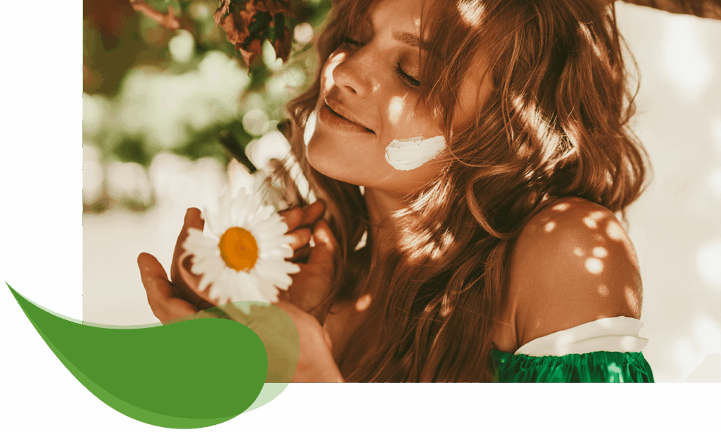 Junge Frau mit langem, welligem Haar genießt den Moment mit geschlossenen Augen unter einem Baum, eine weiße Kamillenblüte in der Hand und Kamillencreme auf der Wange – Symbol für natürliche Hautpflege und Entspannung.