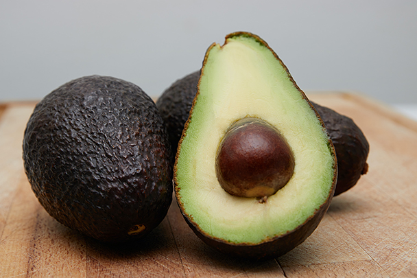 Zwei ganze Avocados und eine aufgeschnittene Hälfte mit Kern auf einem Holzbrett – fruchtige Quelle für pflegendes Avocadoöl
