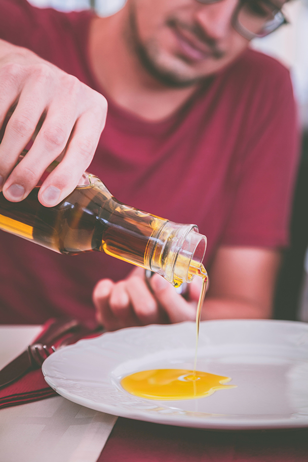 Person gießt goldenes Öl aus einer Glasflasche auf einen Teller – symbolische Darstellung für die ölige Form von Vitamin E Acetate (Tocopheryl Acetate)