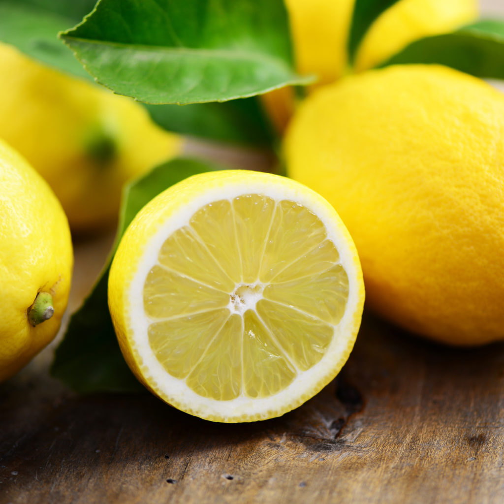 Frisch aufgeschnittene Zitrone mit saftigem Fruchtfleisch, umgeben von ganzen Zitronen und grünen Blättern – Quelle für Citrus Limon Peel Oil mit klärender Wirkung