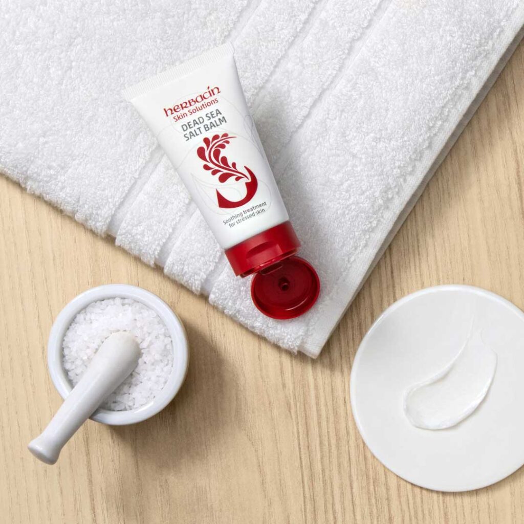 Herbacin Dead Sea Salt Balm auf einem Handtuch mit Meersalzschale und Cremeprobe – beruhigende Pflege für gestresste Haut.