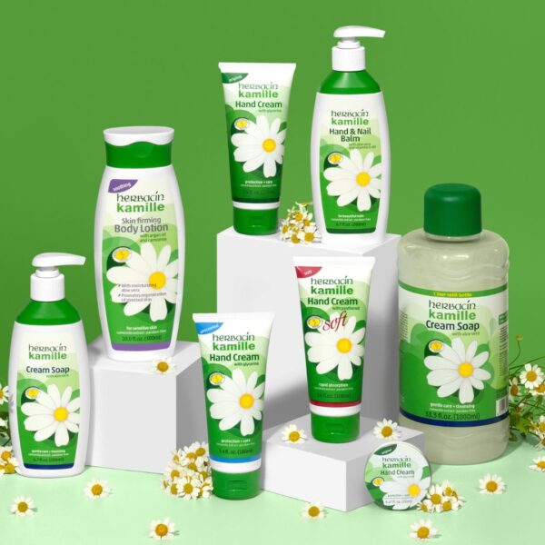 Vollständige Herbacin Kamille Pflegeserie mit Handcreme, Body Lotion, Seife und Nagelbalsam dekorativ auf weißen Podesten mit Kamillenblüten arrangiert.