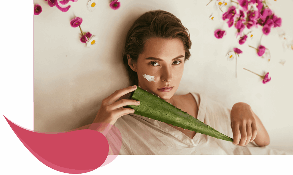 Junge Frau liegt entspannt zwischen Rosenblättern und Kamillenblüten, hält ein großes Aloe-Vera-Blatt in der Hand, Creme auf der Wange sichtbar.