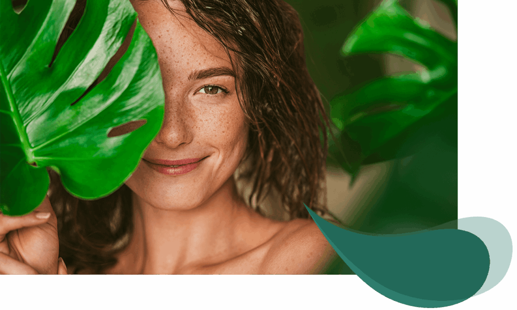 Junge Frau mit Sommersprossen lächelt entspannt hinter einem großen grünen Blatt hervor, natürliche Umgebung, frisches Aussehen.