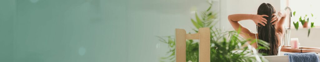 Frau mit langen dunklen Haaren entspannt sich in der Badewanne, Hände im Haar, umgeben von Pflanzen und weichem Tageslicht, frische und natürliche Atmosphäre.