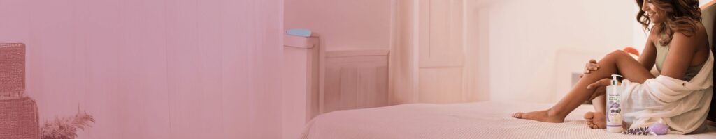 Junge Frau sitzt lächelnd in entspannter Atmosphäre auf einem Bett, trägt ein Handtuch um den Körper, neben ihr ein Lavendelprodukt aus der Herbacin Wellness-Serie.