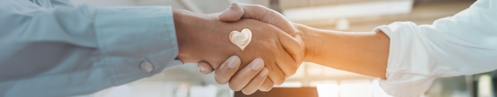Nahaufnahme eines Handschlags zwischen zwei Personen, auf einer Hand ein Herz aus Creme – Symbol für Partnerschaft, Vertrauen und Pflege.