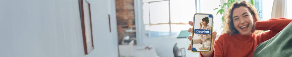 Fröhliche junge Frau hält ein Smartphone mit einer Gewinnspielaktion von Herbacin in die Kamera – Symbol für Freude und Teilnahme an Aktionen.