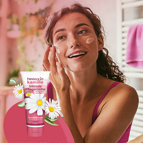 Lächelnde Frau trägt vor dem Spiegel die Herbacin Kamille Intensiv Pflegecreme auf ihr Gesicht auf – im Vordergrund die Creme-Tube mit Kamillenblüten und Vitamin E, ideal für empfindliche Haut.