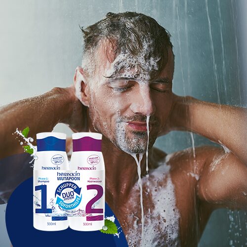 Mann duscht und wäscht sich die Haare mit Shampoo, Schaum läuft über sein Gesicht; im Vordergrund die zwei Phasenprodukte der Herbacin Wutapoon Anti-Schuppen-Serie – wirksame Pflege gegen Schuppen und für gesunde Kopfhaut.