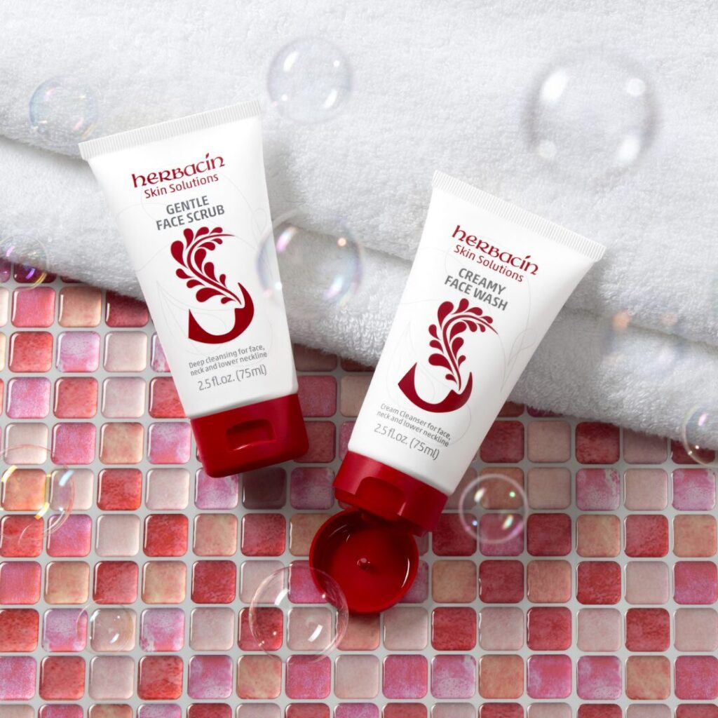 Herbacin Skin Solutions Gentle Face Scrub und Creamy Face Wash auf rosa Mosaikfliesen mit Handtuch und Seifenblasen im Hintergrund.