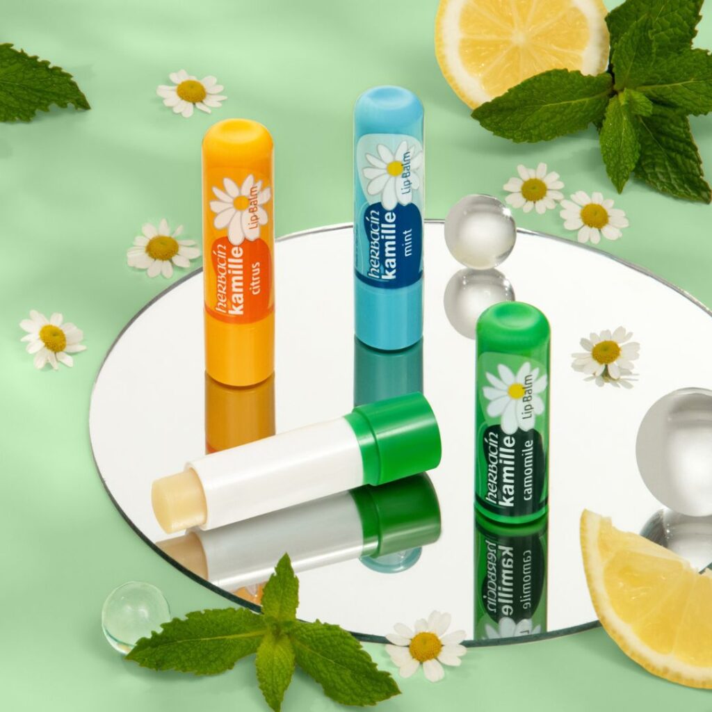 Drei geschlossene und ein geöffneter Herbacin Kamille Lippenpflegestift in den Sorten Citrus, Mint und Original auf einem Spiegel mit Minze, Kamillenblüten und Zitronenscheiben dekoriert.