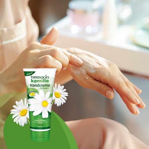 Person cremt sich im Sitzen die Hände mit Herbacin Kamille Handcreme ein, im Vordergrund Verpackung mit Kamillenblüten abgebildet