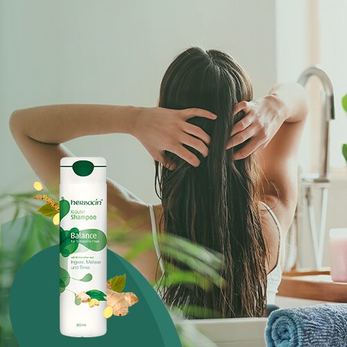 Frau sitzt mit dem Rücken zur Kamera und fährt sich durch das nasse Haar; im Vordergrund steht das Herbacin Kräuter Shampoo mit Zitronengras & Ingwer – Symbol für erfrischende Reinigung und natürliche Pflege.
