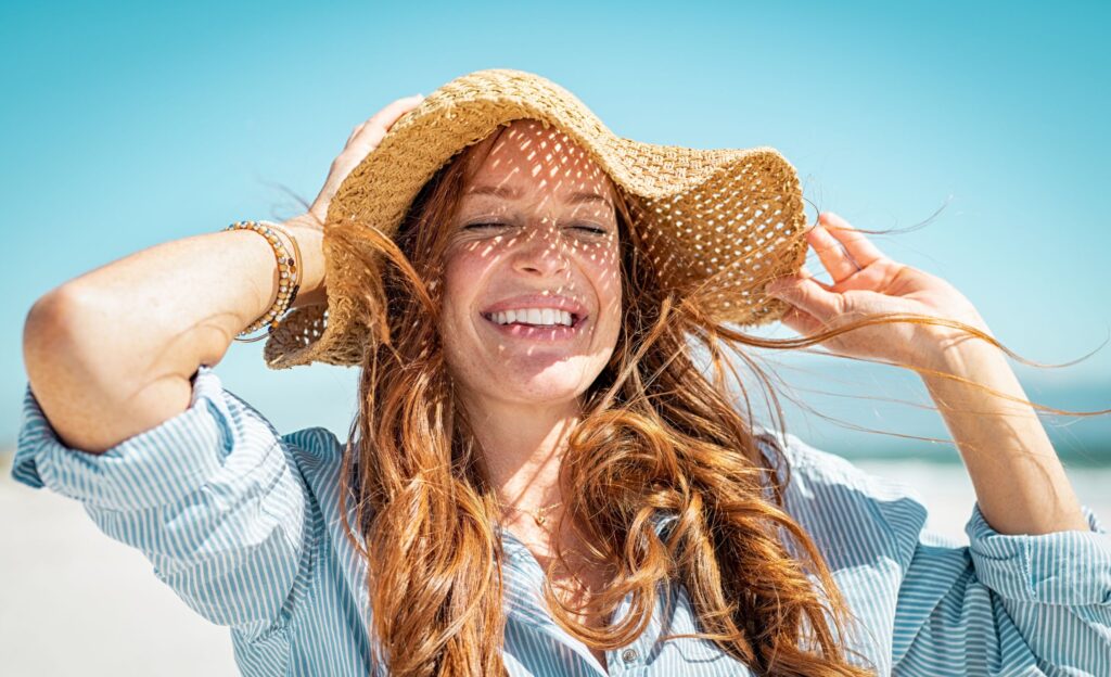 Auf dem Bild ist eine Frau am Strand zu sehen. Sie hat rote lange Haare, trägt einen Strohhut und lächelt.